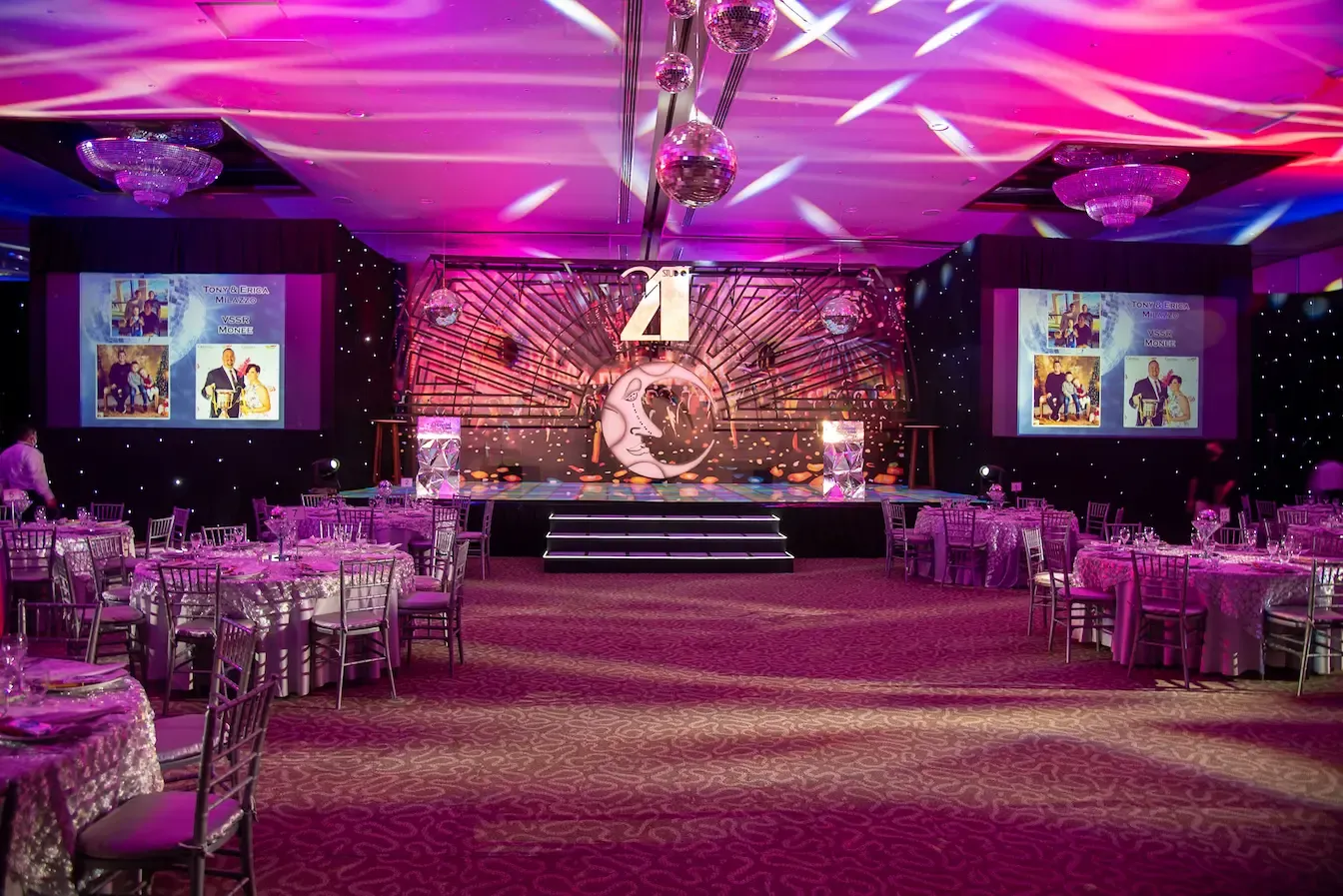 A decorated event ballroom with a themed stage backdrop, disco balls and round banquet tables